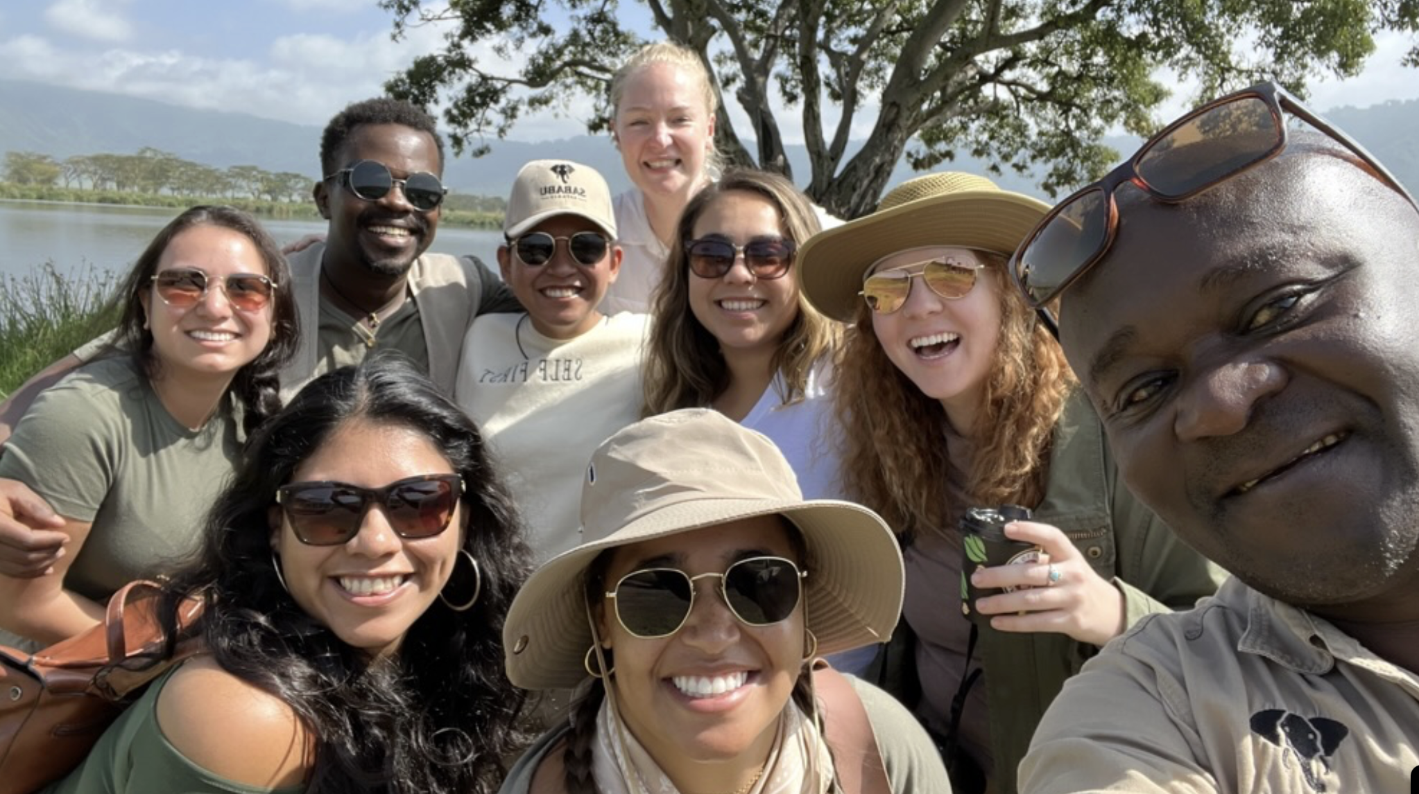 Group of ten smiling people outdoors near water, wearing sunglasses and hats, taking a selfie together with a mountain and tree in the background.