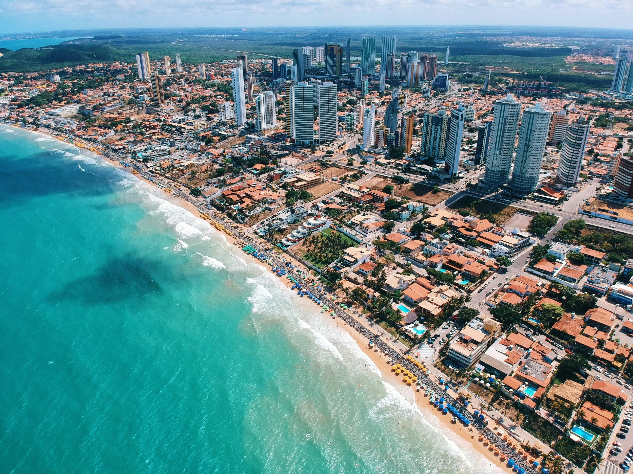 Aerial view of a beachfront city with high-rise buildings, a sandy beach with umbrellas, and turquoise ocean water.