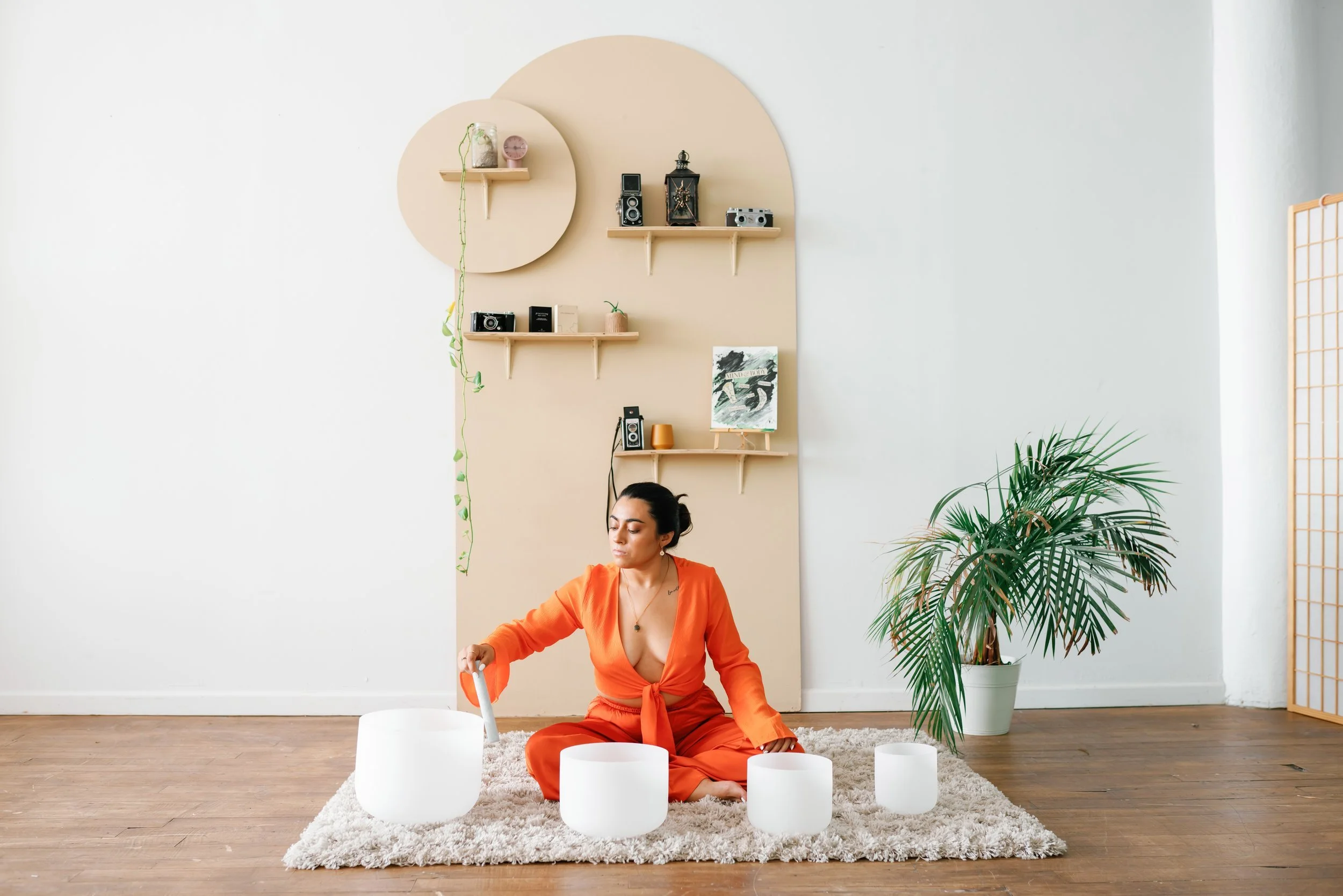 A woman in an orange outfit sits on a beige shaggy rug playing crystal singing bowls in a minimalist room with white walls, wooden flooring, a large potted plant, and decorative shelves with cameras and artwork.