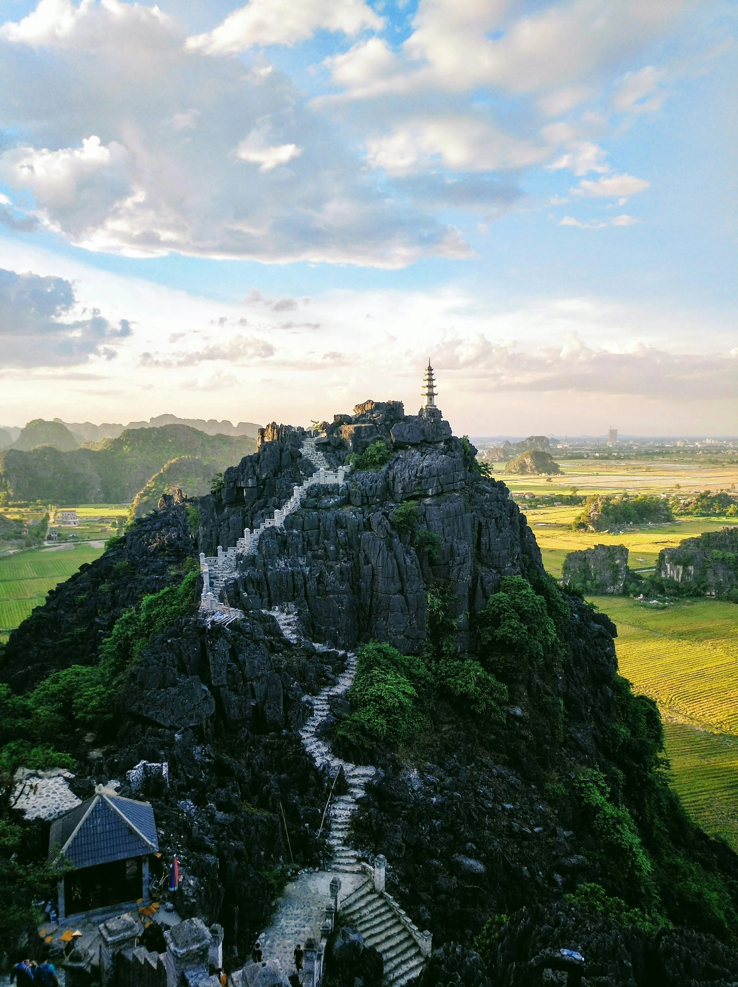 A mountain with a staircase leading to a pagoda at the top, surrounded by green vegetation and overlooking fields and distant hills.