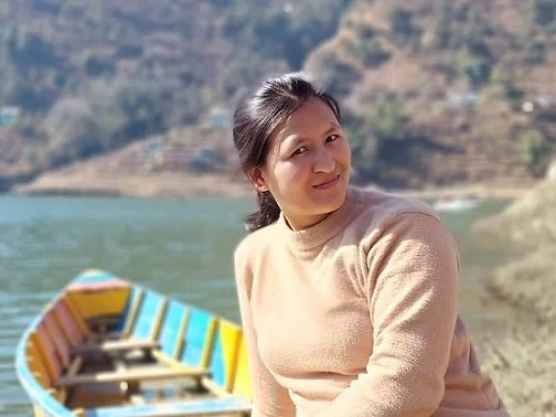 A woman sitting by a colorful boat on a river with hills in the background.