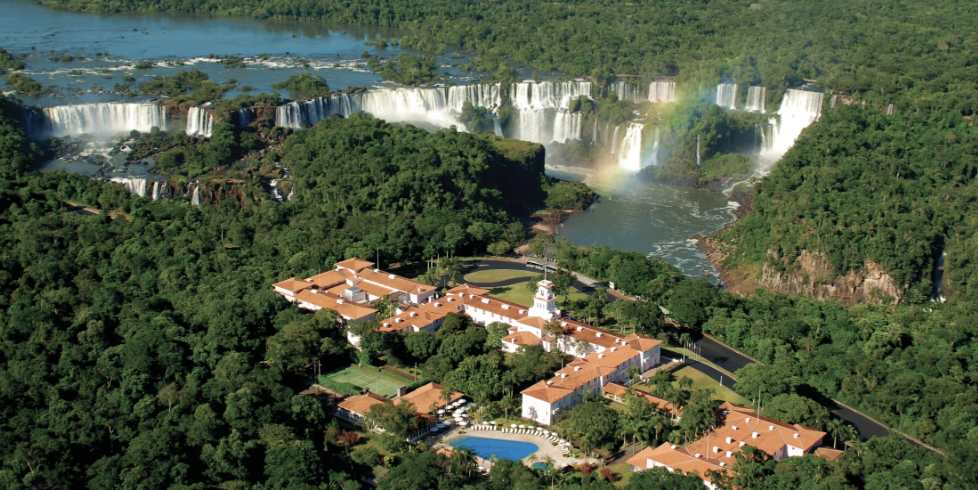 Aerial view of Iguazu Falls surrounded by lush green forest and a resort with white buildings and red roofs in the foreground.