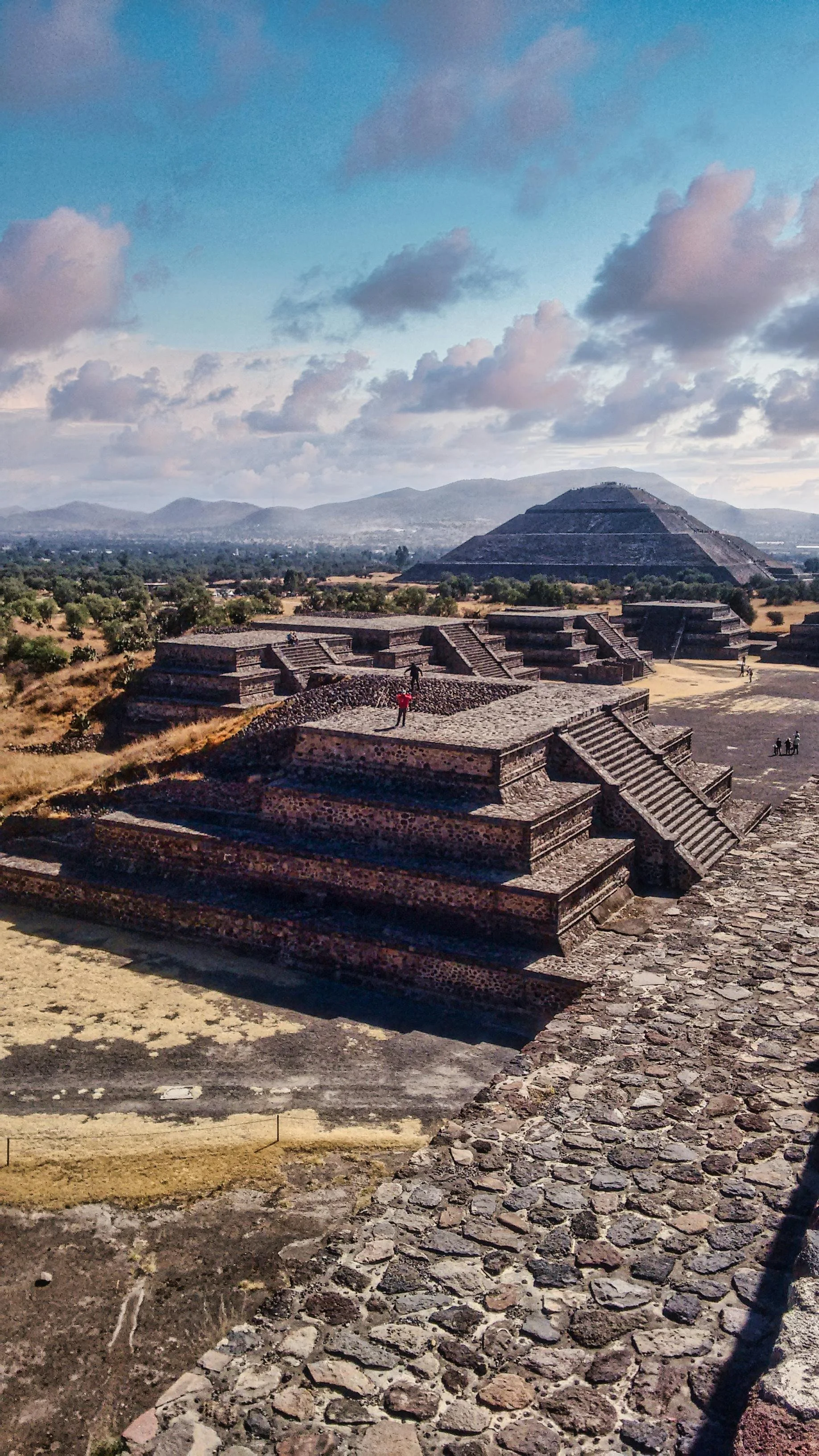 Ancient stepped pyramid at Teotihuacan with smaller pyramids and stairs, cloudy sky, distant mountains, and a few people exploring the site.