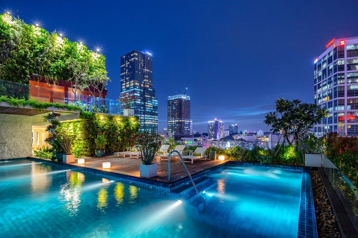 Rooftop swimming pool at night with city skyline, lit plants, and lounge chairs.