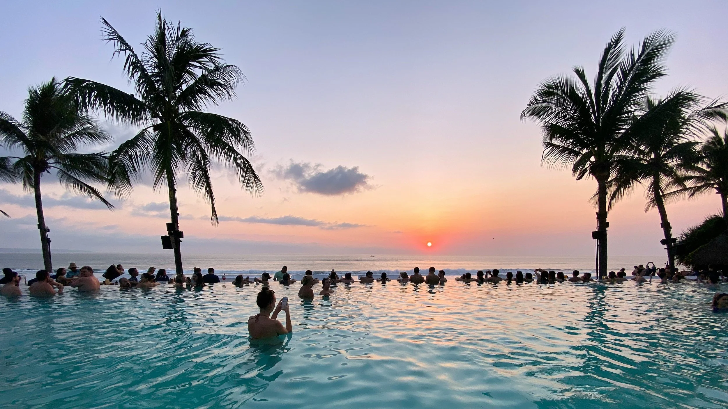 People swimming and relaxing in a pool during sunset with palm trees and ocean in the background.