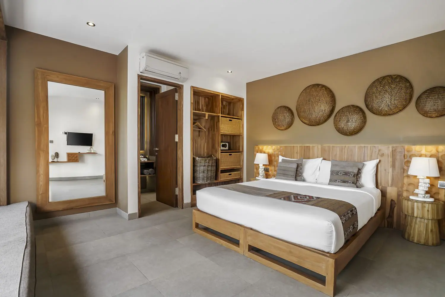 A hotel room with a large bed, wooden headboard, and decorative woven baskets on the wall. Two bedside lamps, a wooden shelving unit, and a mirror are visible. An open door leads to a bathroom area, and a large mirror reflects a second room with a TV.