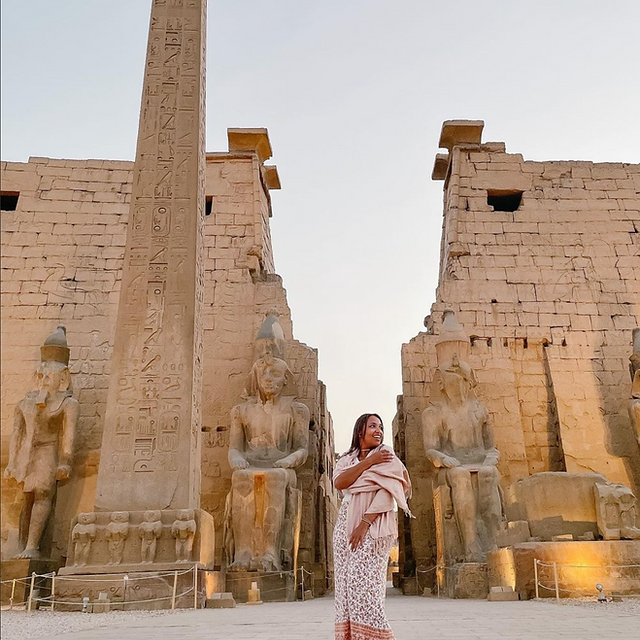 Woman in patterned dress standing amidst ancient Egyptian statues and ruins.