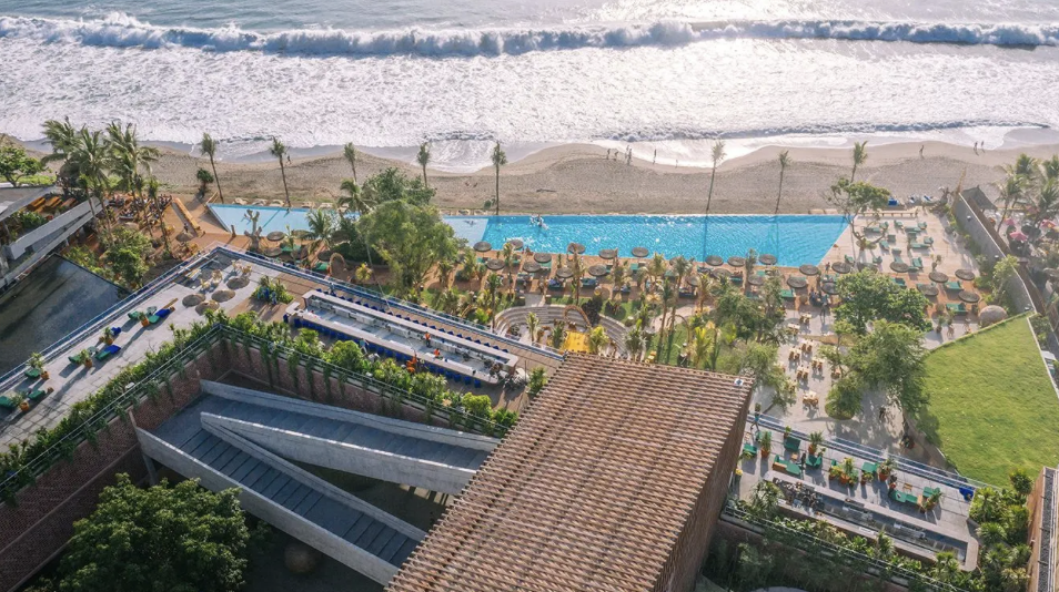 Aerial view of a beachfront resort with a pool, lounge chairs, and palm trees overlooking the ocean.