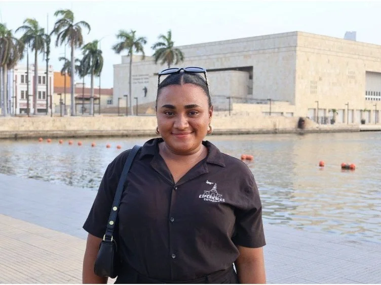 Female tour guide A woman standing outdoors near a body of water with palm trees and a large modern building in the background.