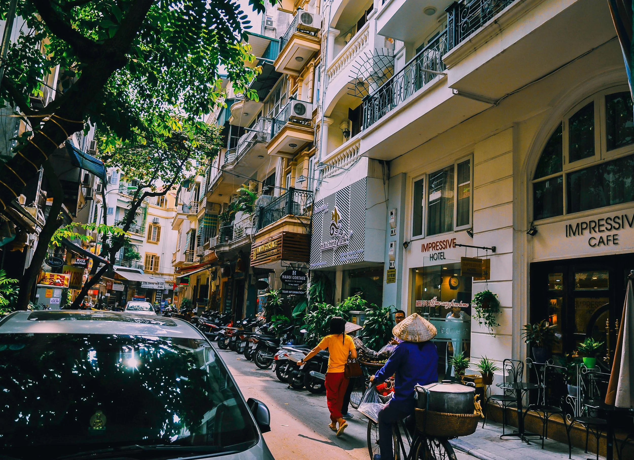 A busy city street in Vietnam with parked motorbikes, pedestrians, and a cyclist. Multi-story buildings with balconies and shops line the street, with trees providing shade.