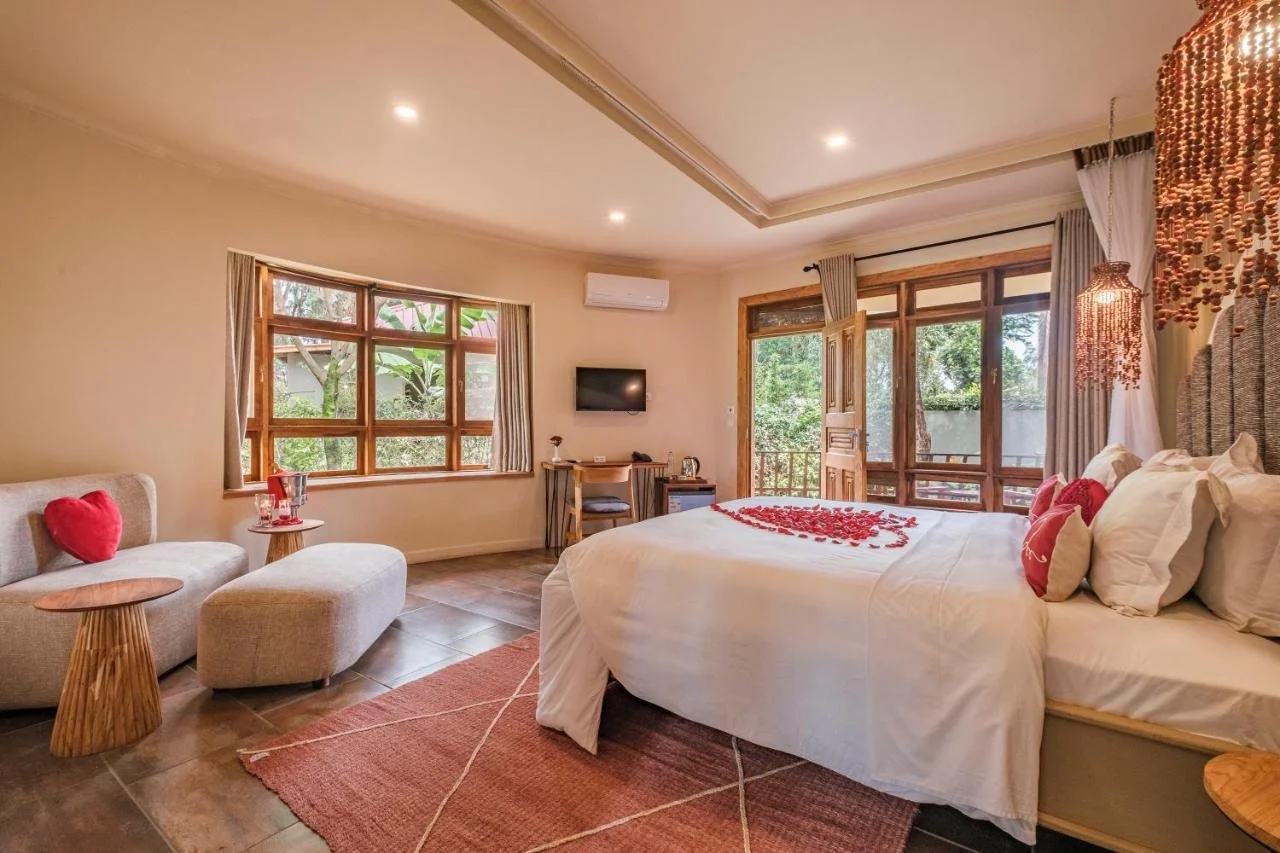 A cozy hotel room with a large bed decorated with rose petals, a sitting area with a beige sofa and small tables, and a view of greenery outside through large windows and a balcony.