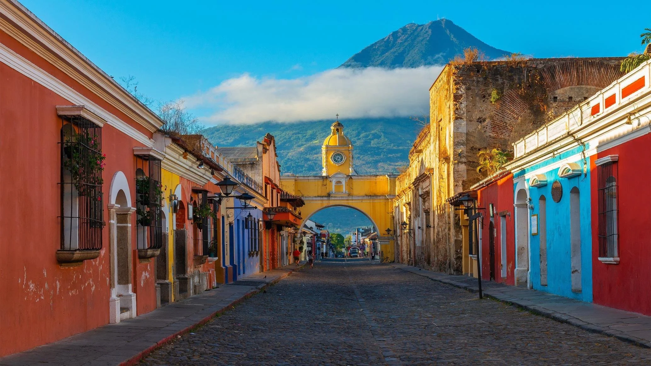 Colorful colonial-style buildings along a cobblestone street, with a yellow clock tower archway, a mountain in the background, and a volcano partially covered by clouds.