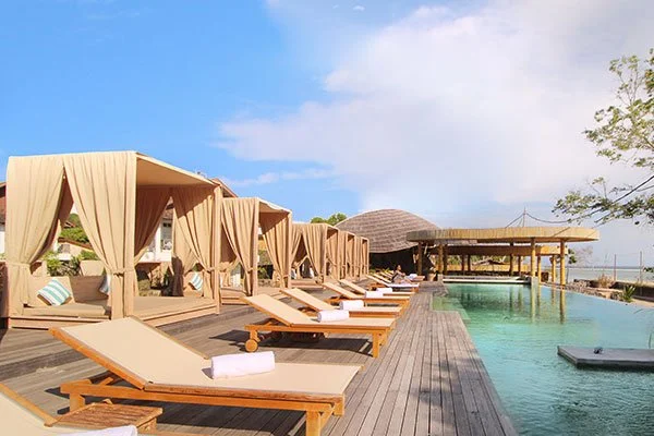 Poolside lounge chairs with beige curtains and white towels beside an infinity pool under a partly cloudy sky at a resort.
