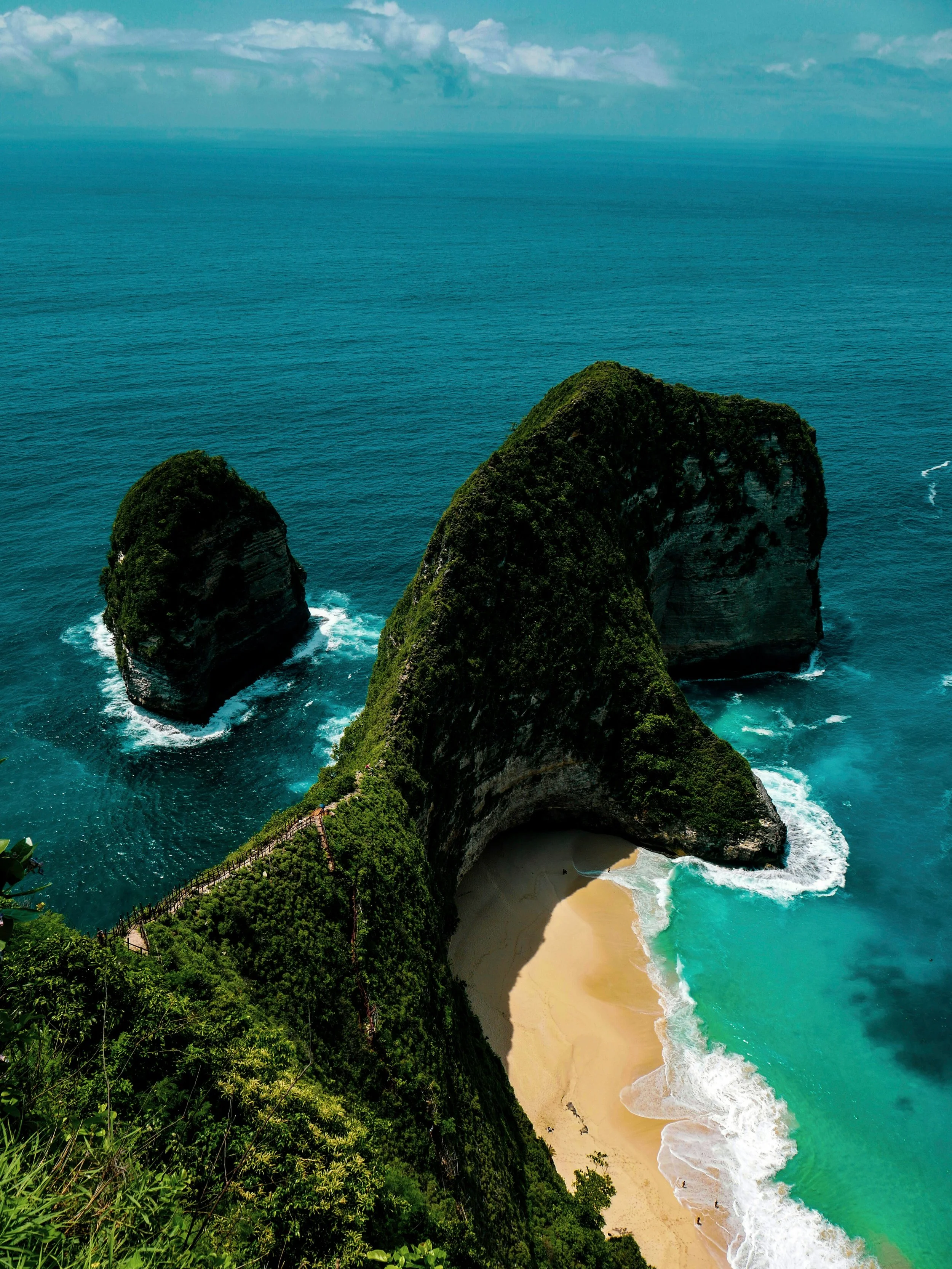 Aerial view of a beach with large green cliffs and rock formations surrounded by blue ocean water.