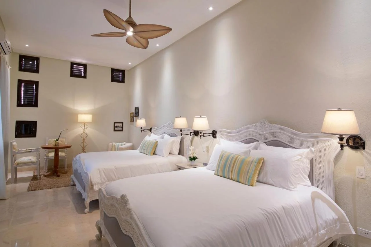 A bedroom with two white beds, each with striped pillows, wall-mounted lamps, a small round table with a chair, a floor lamp, and windows with dark shutters.