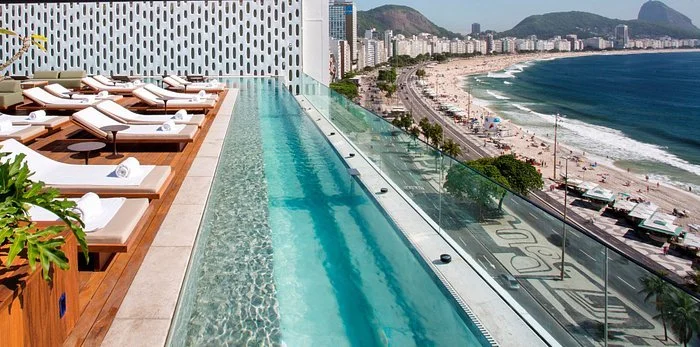 Luxury rooftop infinity pool overlooking a beach with high-rise buildings, mountains, and ocean in the background.