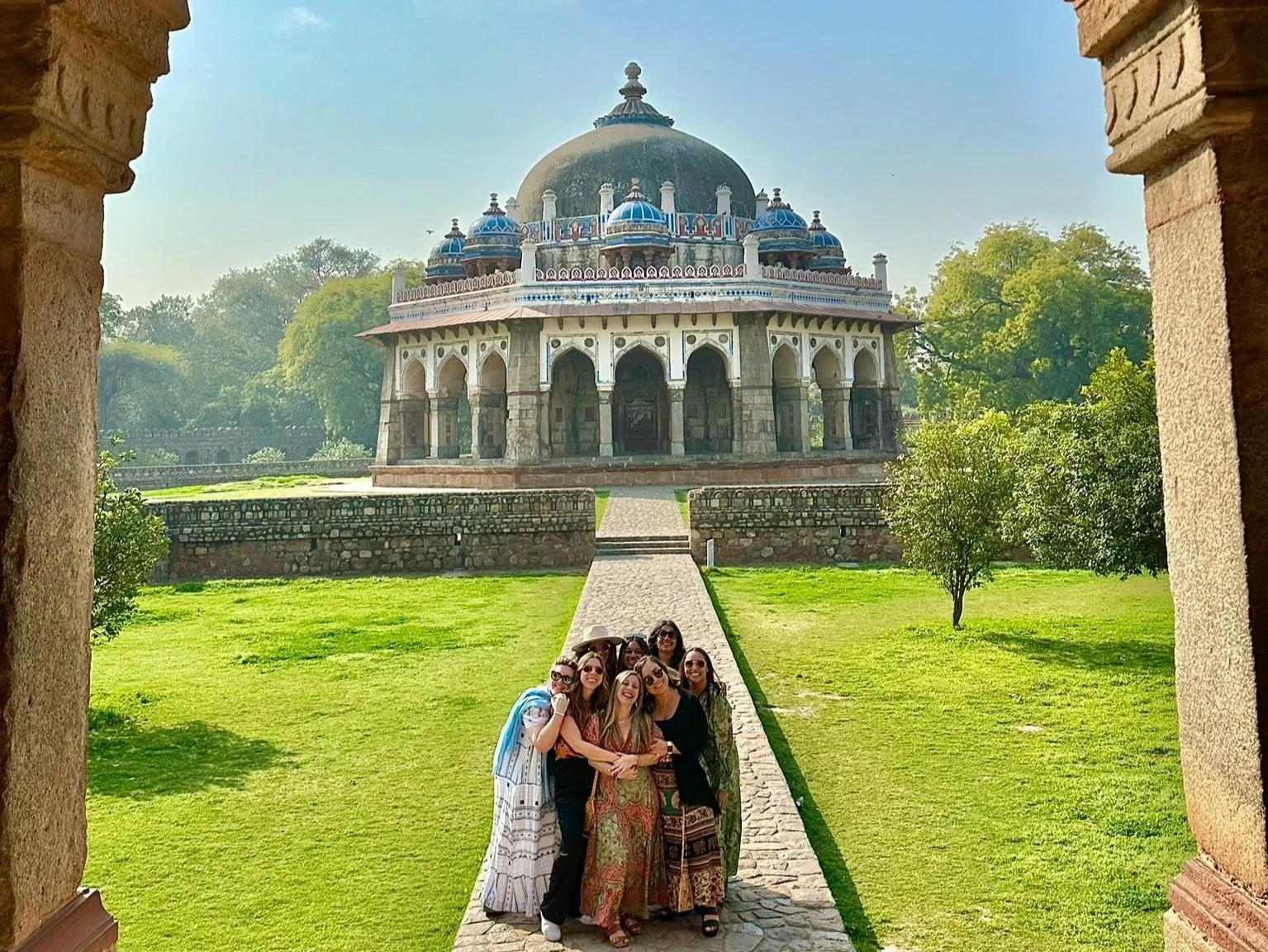 A group of seven women posing together on a stone pathway, in front of a historic white monument with a large central dome and smaller blue domes, surrounded by greenery and trees, under a clear blue sky.