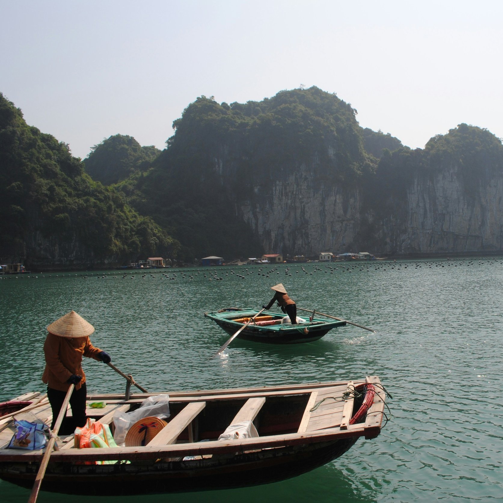 Two individuals wearing traditional Asian conical hats are rowing small boats on calm water with limestone cliffs and small houses in the background.