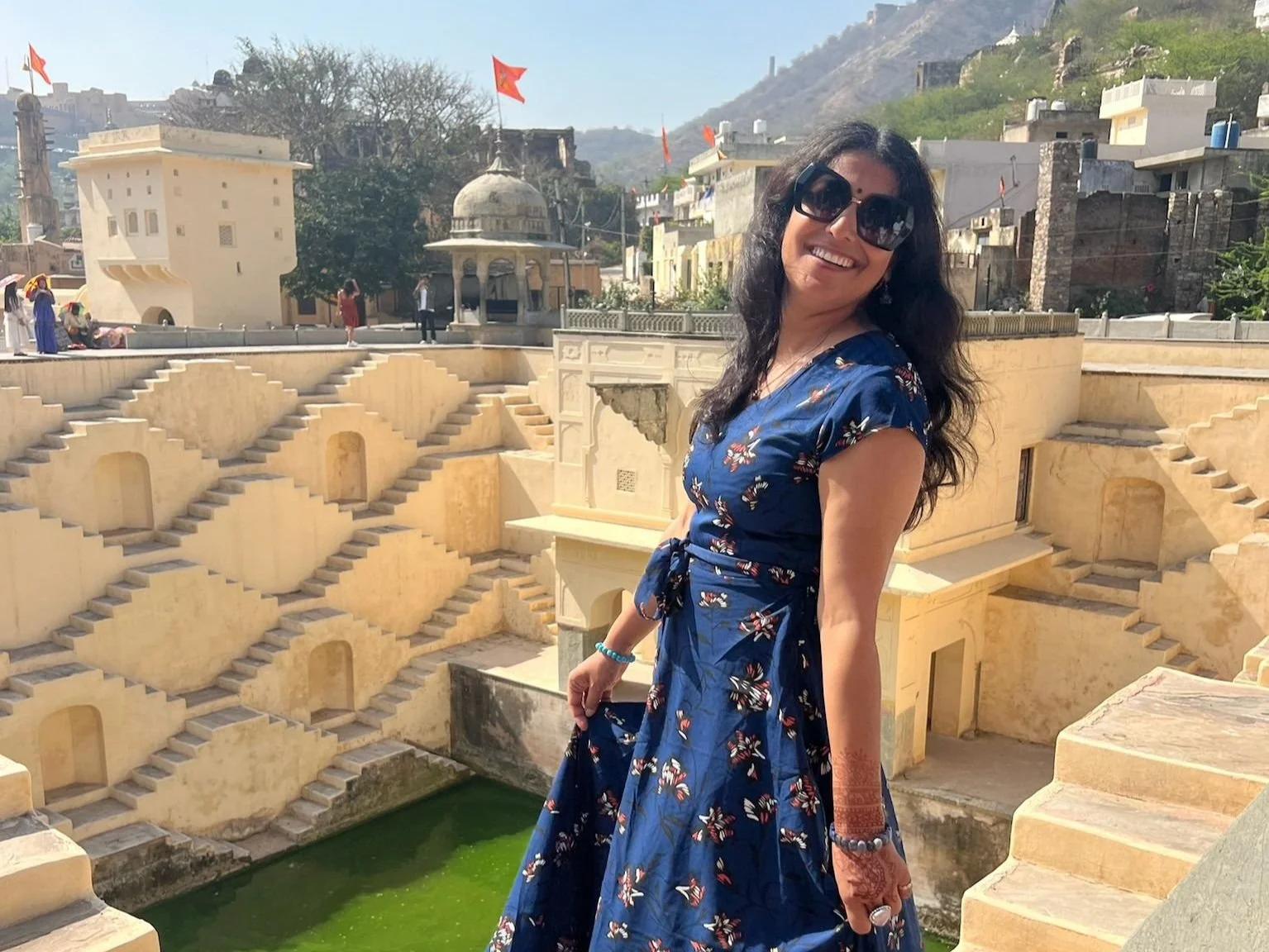 Woman smiling and wearing a blue floral dress and sunglasses near historic stepwell with ornate architecture, stairs, and water, in an outdoor setting with hills and buildings in the background.