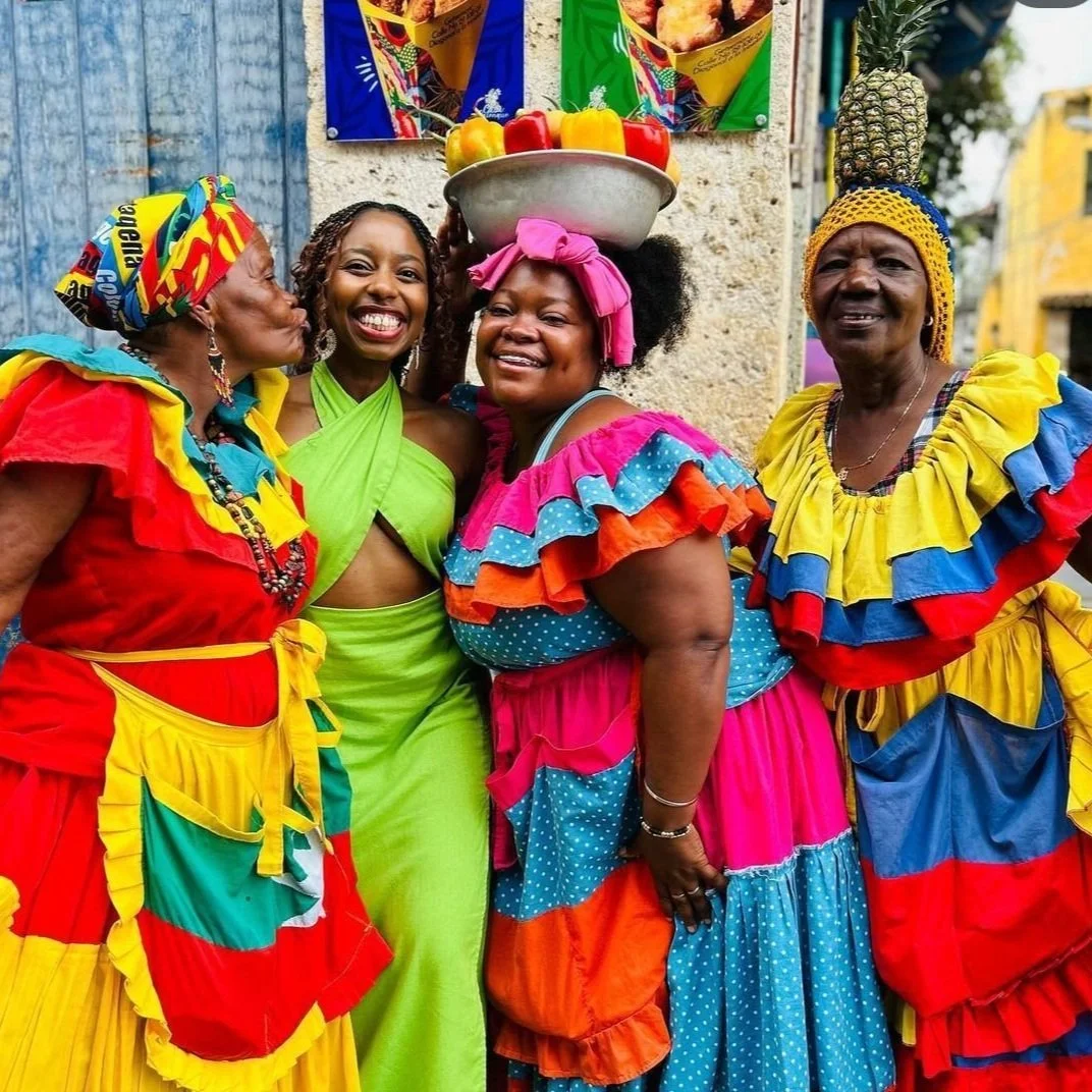 Four women in colorful traditional dresses and accessories smiling and posing together outdoors, with one woman balancing a bowl of fruits on her head.