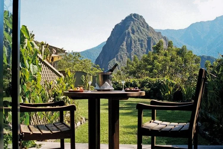 Outdoor patio with a round table set for two, surrounded by two chairs, overlooking a lush green garden and a mountain in the background.