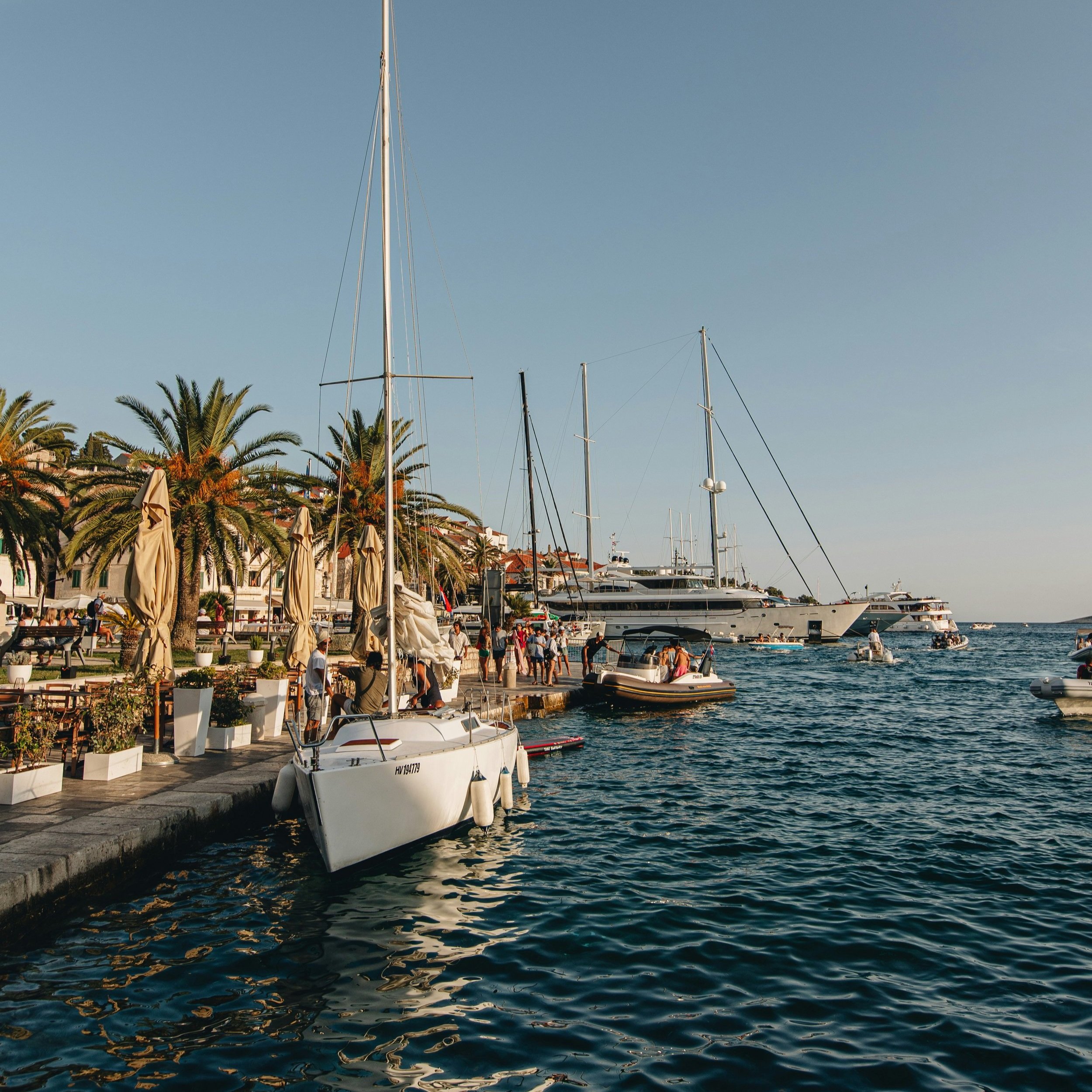Boats docked at a marina with people walking along the waterfront promenade, palm trees, and buildings in the background under a clear sky.