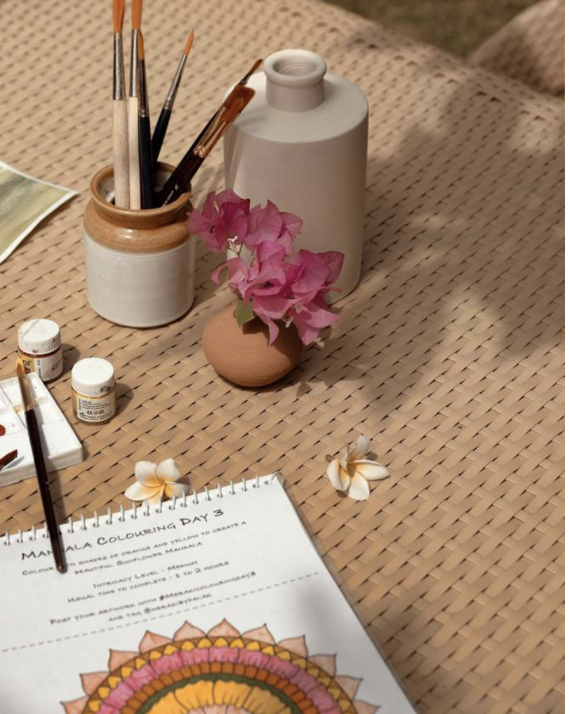A table with a pencil holder containing paintbrushes, a small white bottle, a larger white bottle, a pink flower in a small brown vase, a paint palette, and a coloring book open to a mandala design.