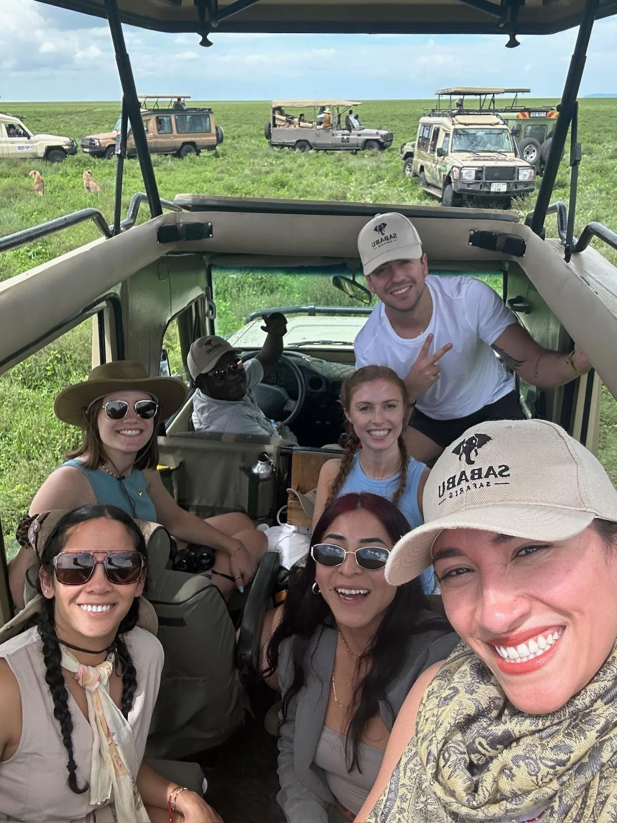 A group of seven people smiling for a selfie inside a safari vehicle with open sides, in a grassy landscape with several safari vehicles and lions in the background.