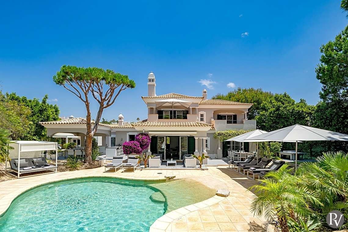 Luxury house with a backyard pool, sun loungers, umbrellas, and lush greenery under a blue sky.