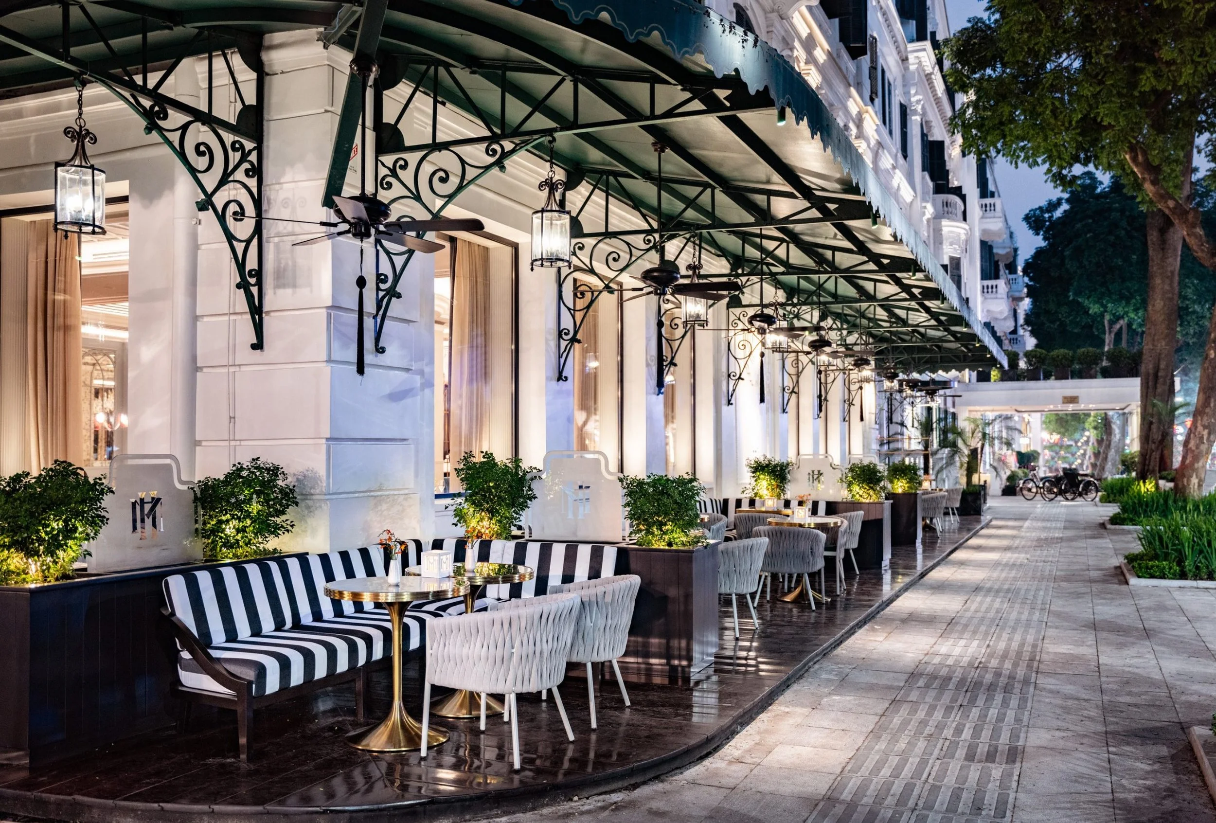 An outdoor restaurant patio with black and white striped seating, small gold tables, green plants, and elegant lighting fixtures under a canopy, with trees and a sidewalk in the background.