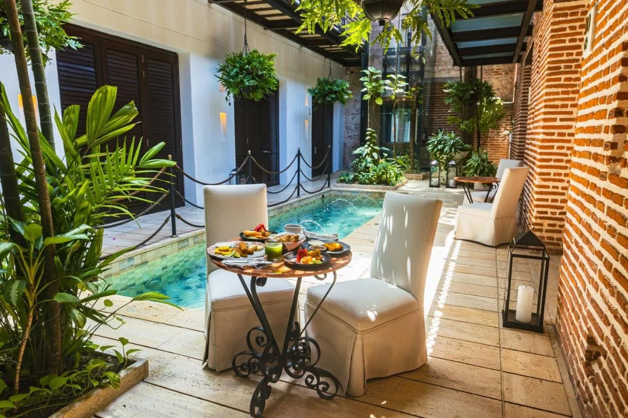 Outdoor patio with a small pool, white chairs, a table with breakfast, green plants, brick and glass walls, and lanterns.