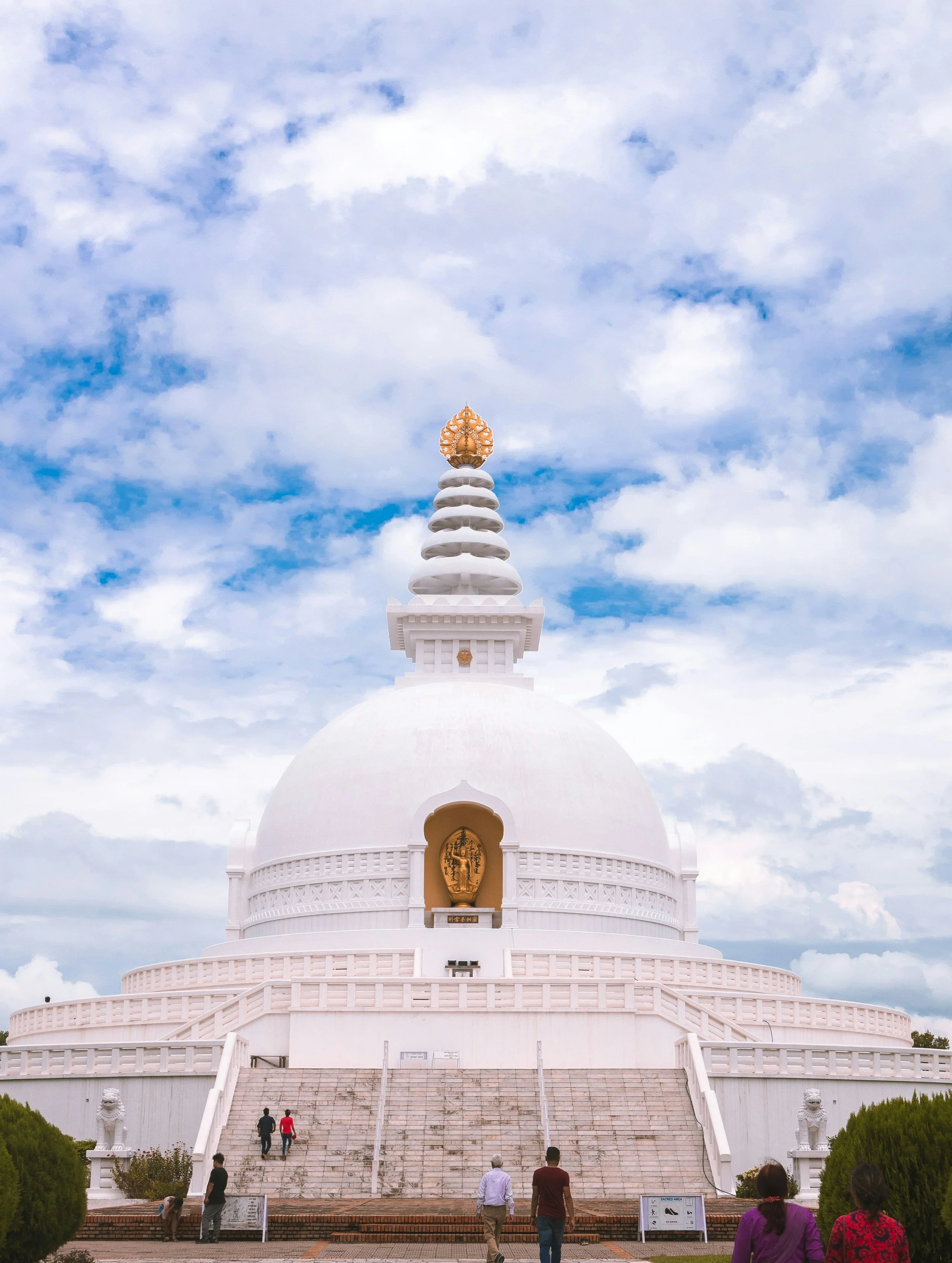 White Buddhist stupa against cloudy sky with stairs leading up to it and people walking nearby.