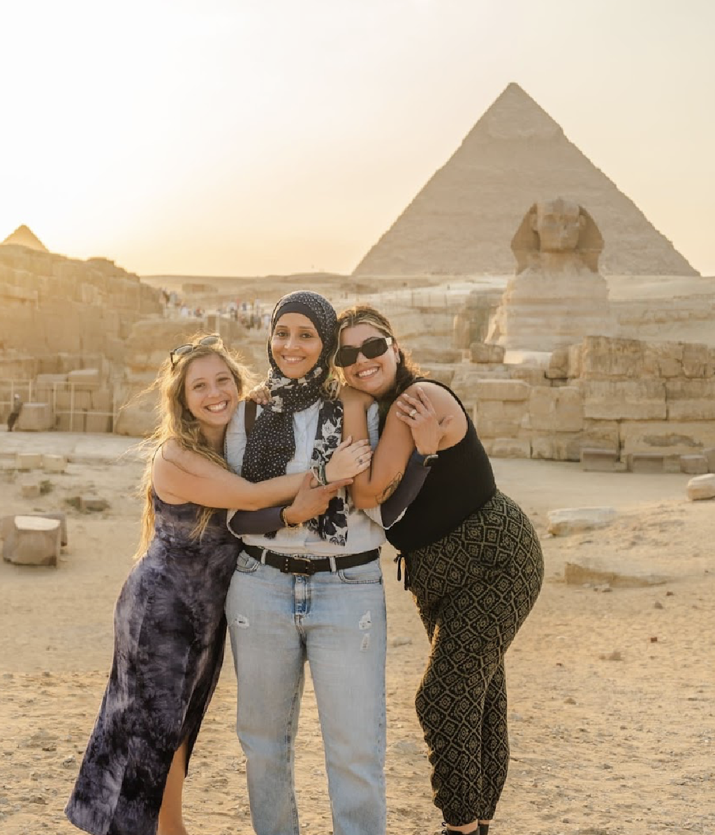 Three women smiling and hugging in front of the Great Pyramid of Giza and a Sphinx statue at sunset in Egypt.