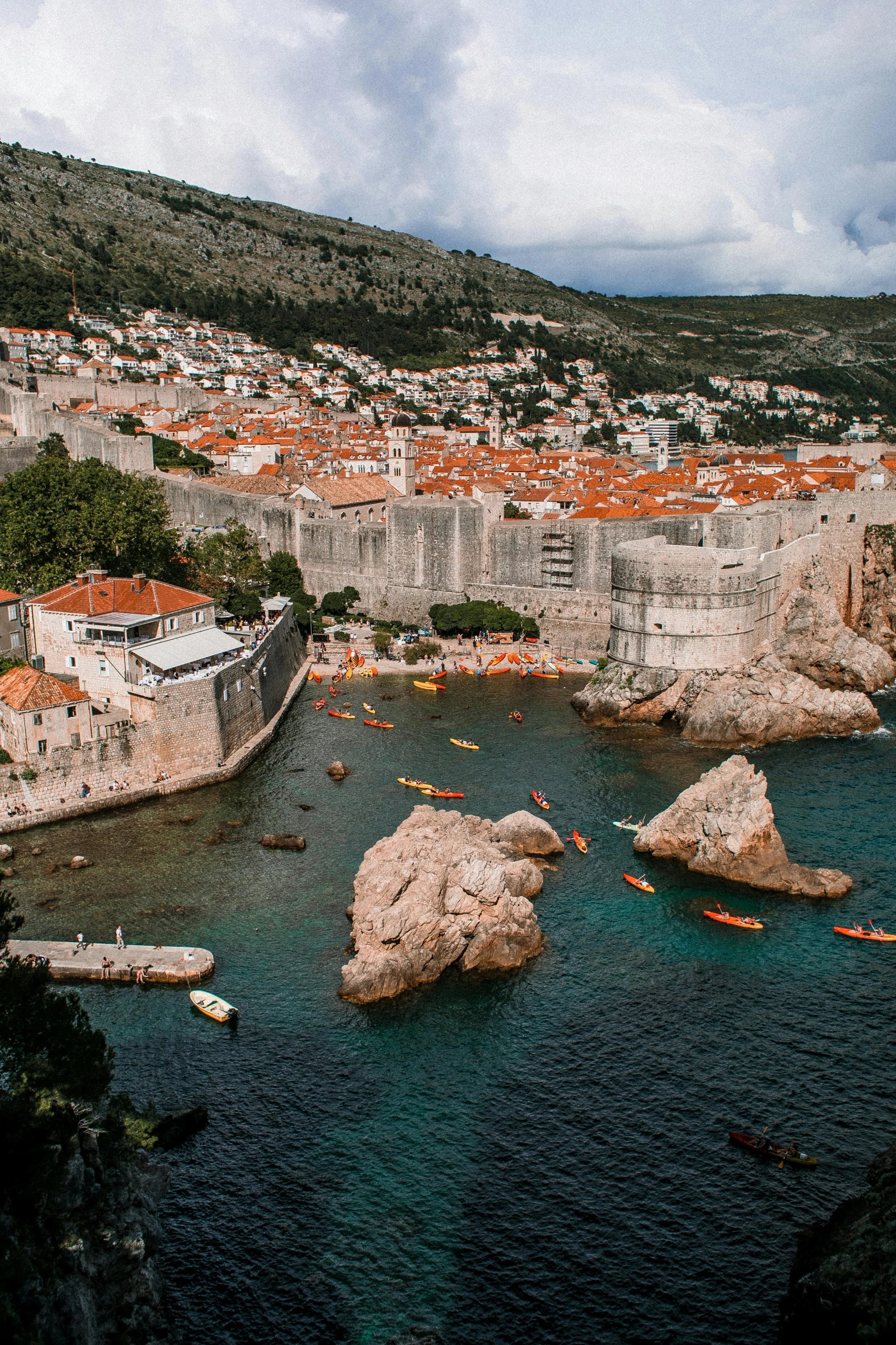 A coastal city with historic stone buildings, red-tiled roofs, a large fortress wall along the water, and rocky rocks in the bay with kayaks and boats.
