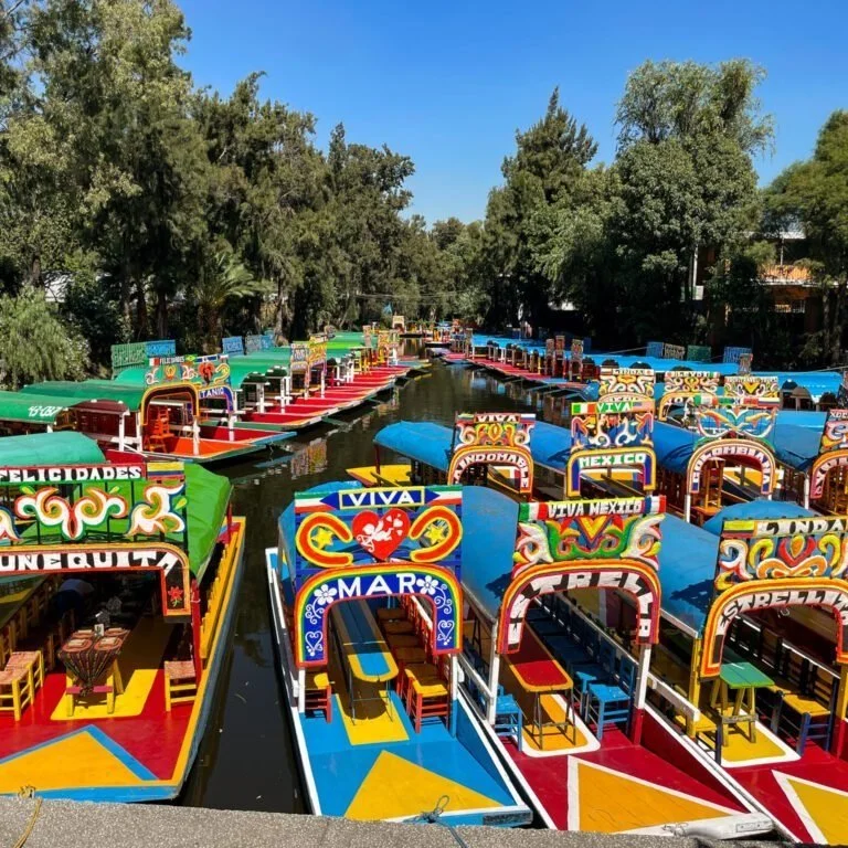 Colorful boats docked along a canal, each decorated with vibrant signs representing different Mexican states and phrases, under a clear blue sky.