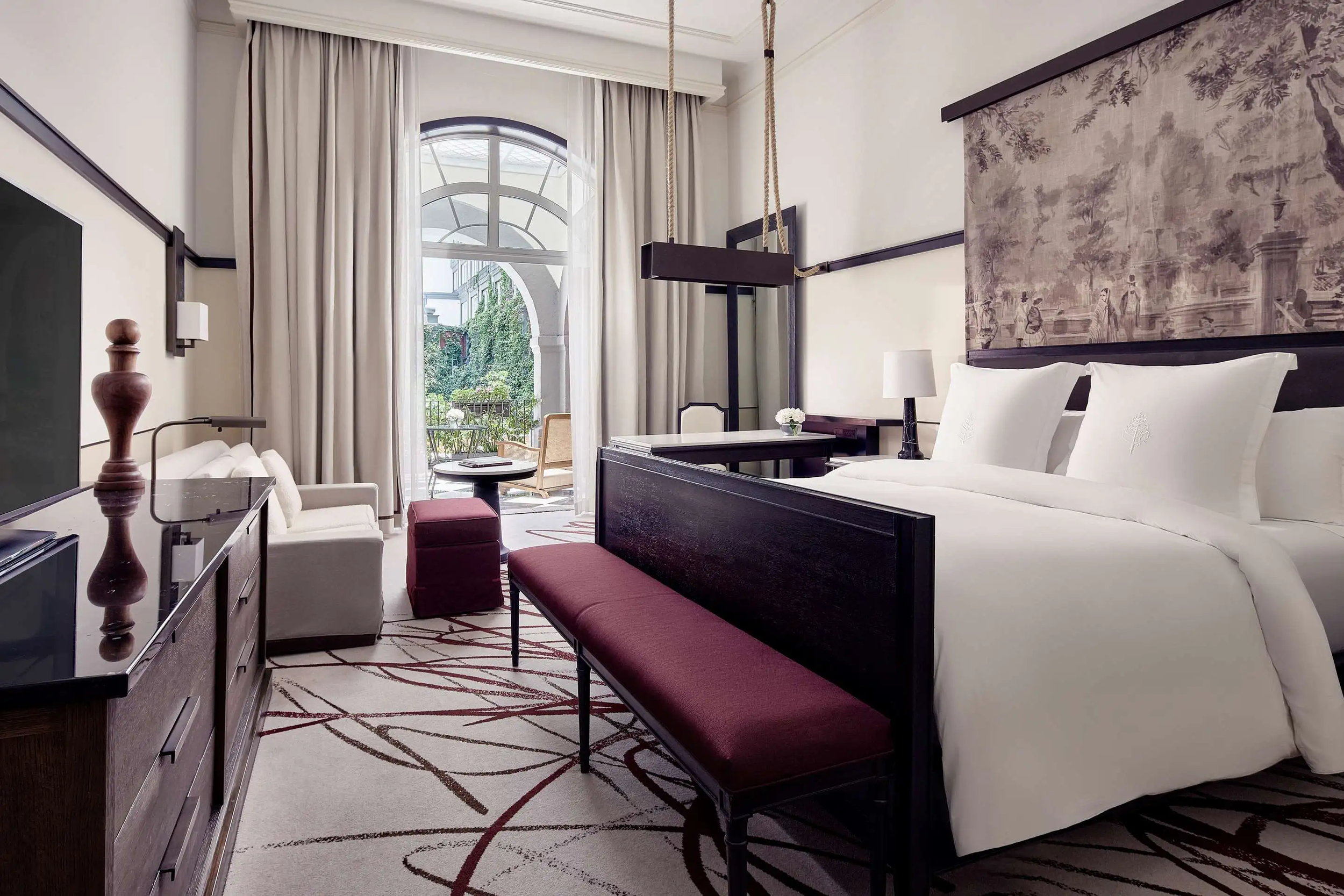 Luxury hotel room with a large bed, white linens, a sitting area with two chairs and a small table, a bench at the foot of the bed, a dresser with a TV, and a balcony with outdoor furniture visible through glass doors.