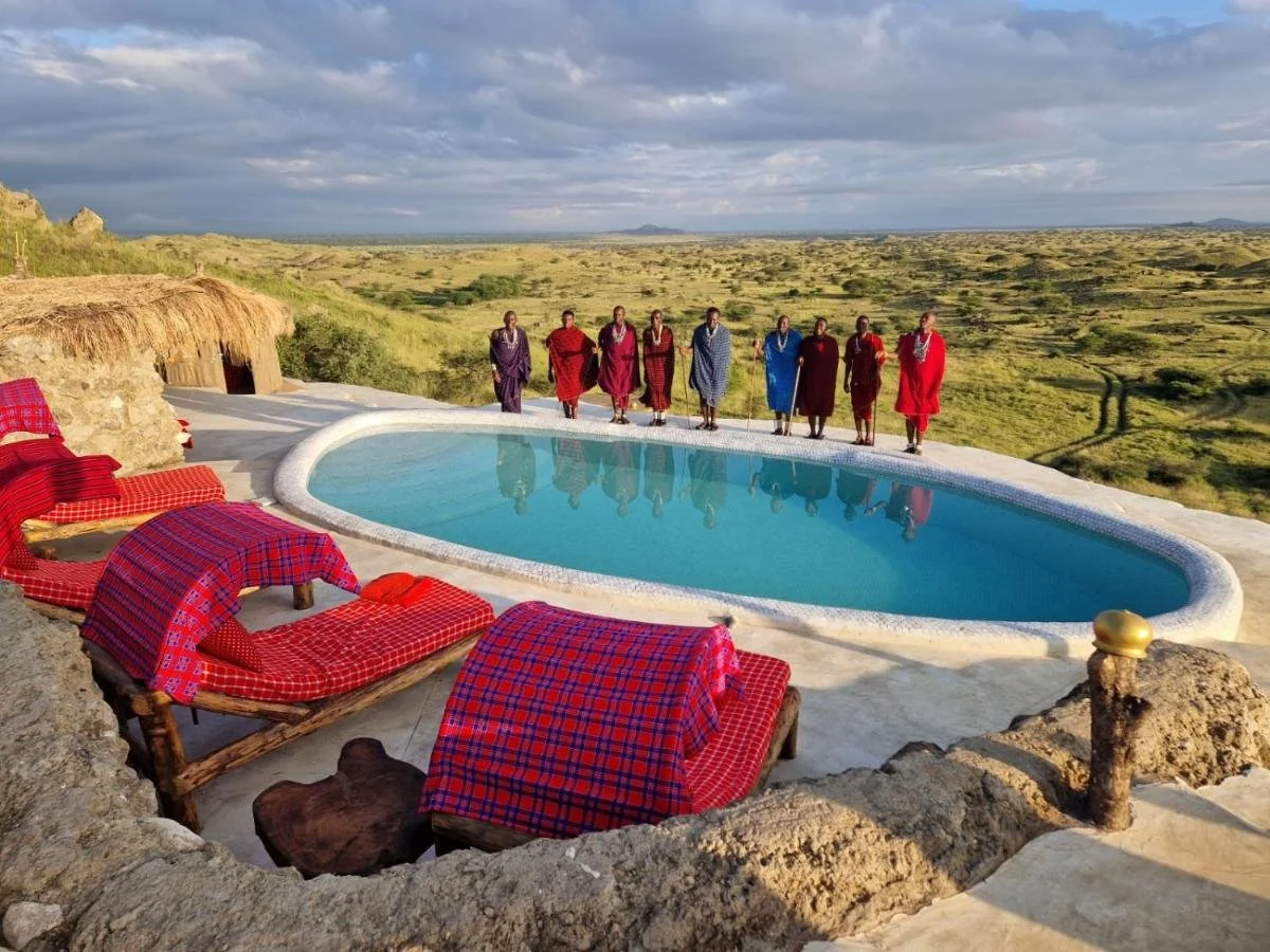 A group of nine Maasai men dressed in traditional red and blue shukas standing by a pool with a view of a grassy plains landscape.