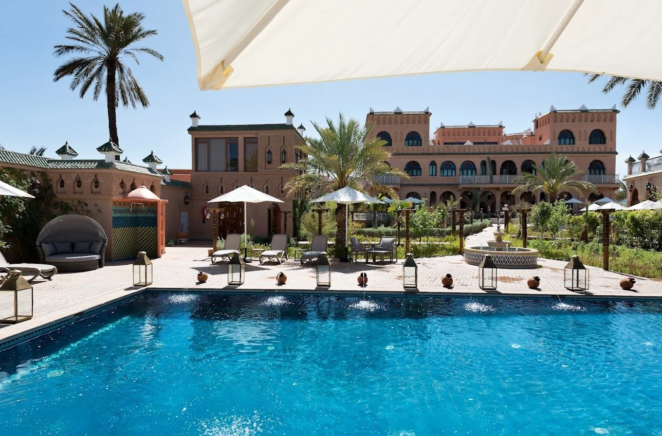 Luxury hotel courtyard with a swimming pool, sun loungers, umbrellas, and palm trees under a clear blue sky.