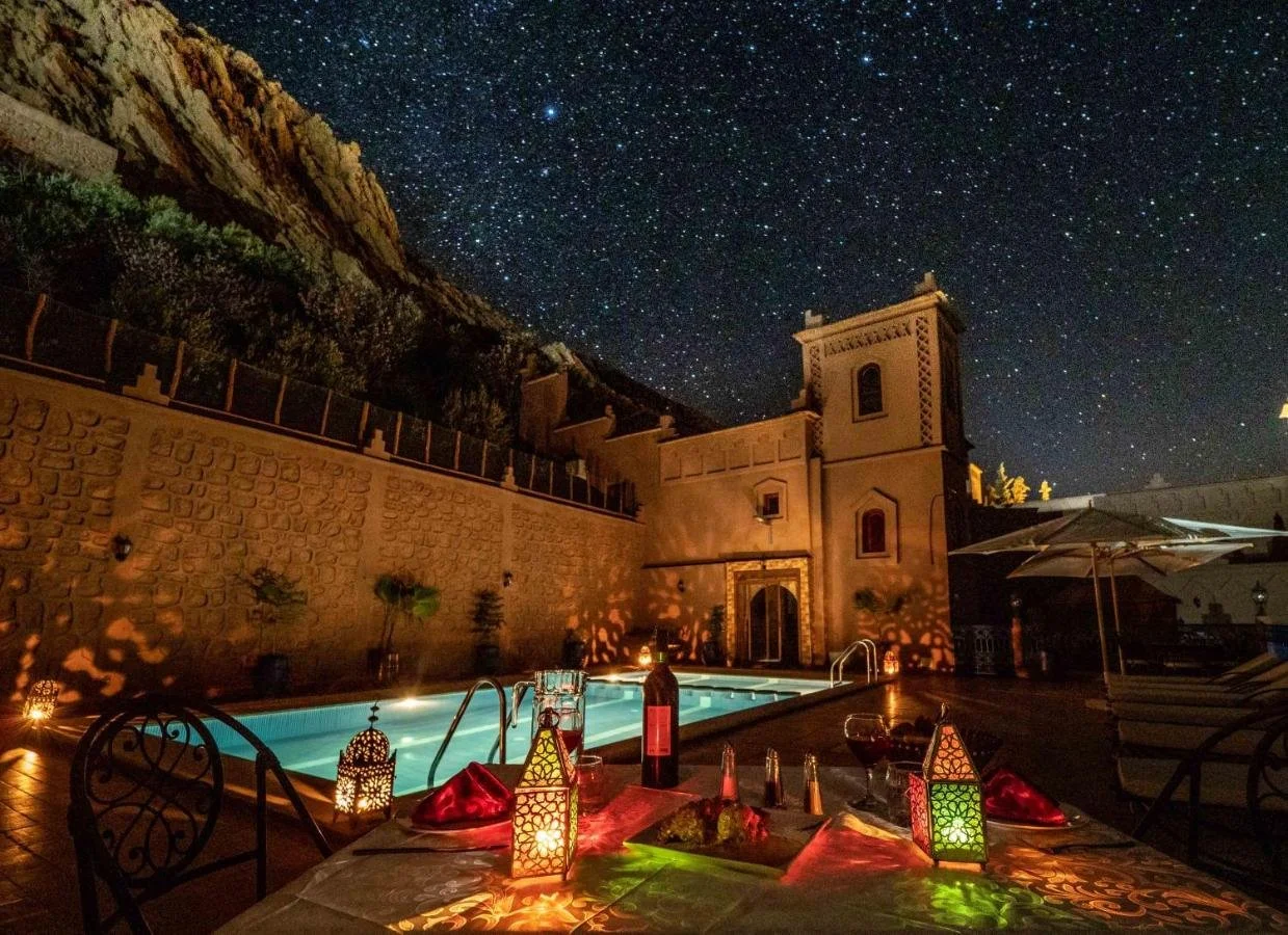 Nighttime outdoor scene of a poolside dinner with table setup, decorative lanterns, and a historic building in the background under a starry sky.