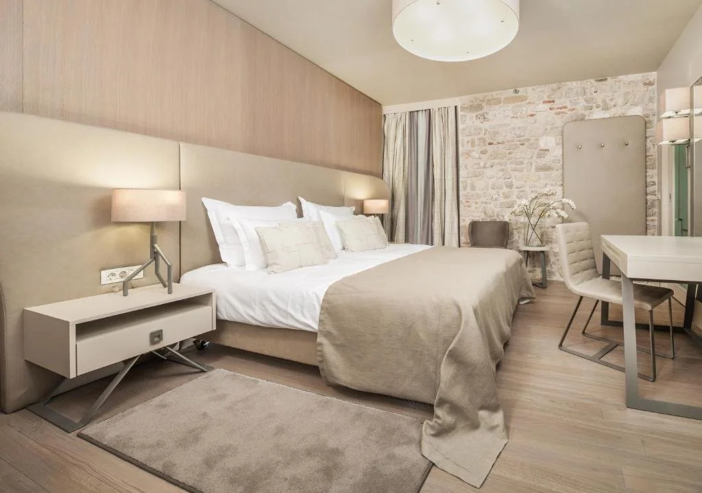 Modern hotel room with a large bed, beige bedding, bedside lamps, a gray chair, a small table with flowers, a desk and chair, and a brick accent wall.