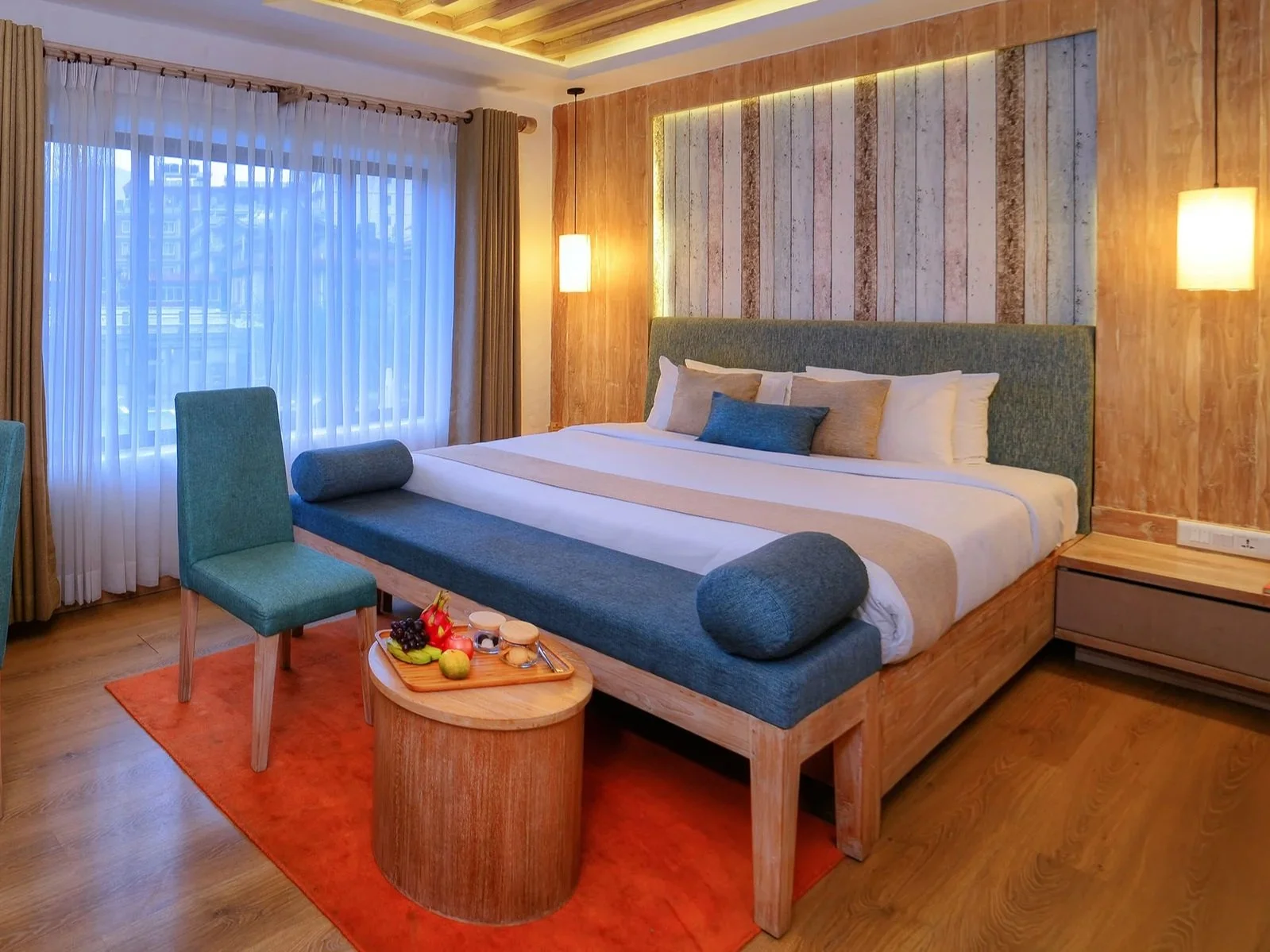 A modern hotel room with a large bed, wooden accents, and a view outside through sheer curtains. There are two teal chairs and a small wooden table with fruit and snacks, and warm lighting.