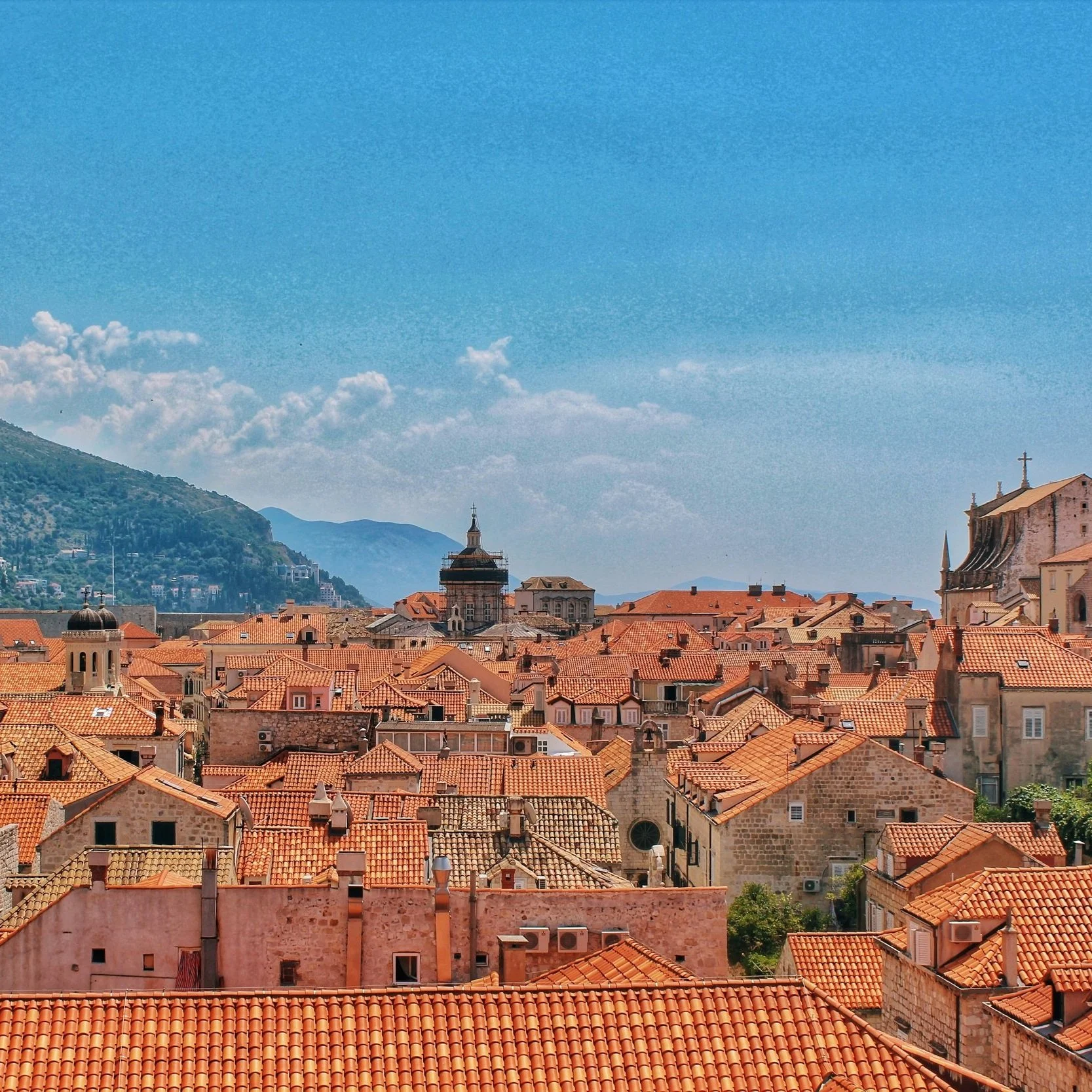 View of a European city with red-tiled roofs, historic buildings, and mountains in the background under a clear blue sky.