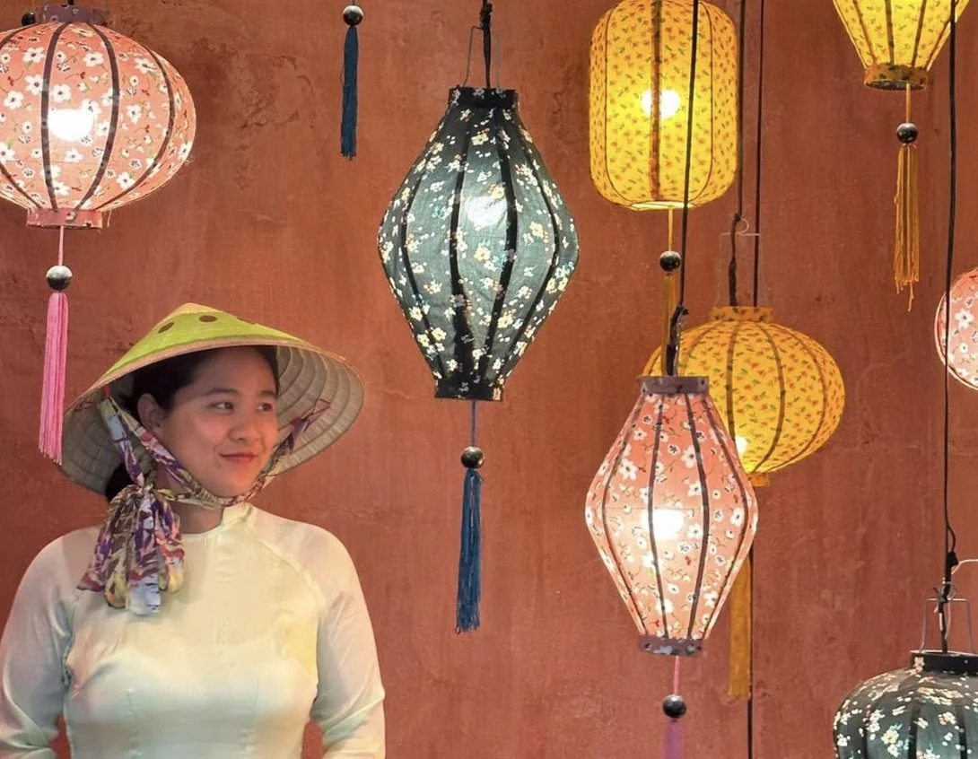 A woman wearing a traditional conical hat with a scarf wrapped around her neck, standing in front of colorful hanging lanterns against a brick wall.