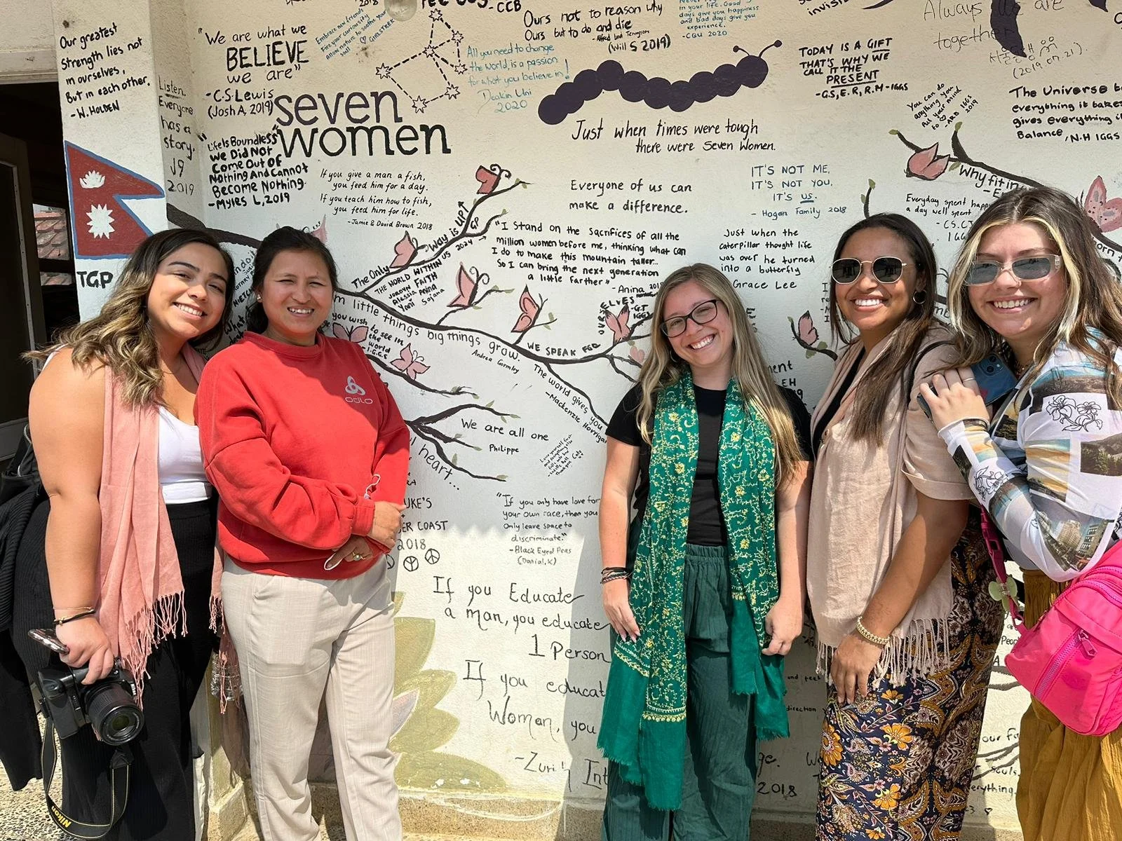 Five women standing outside in front of a large white wall covered with handwritten quotes and drawings. They are smiling and posing for the photo. The wall has musical notes, butterflies, and motivational messages.