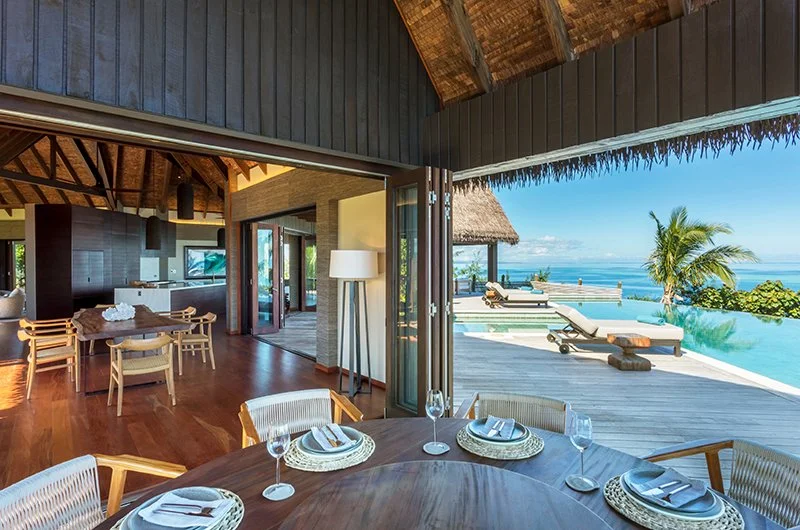 Open-air dining area with round wooden table set with plates, glasses, and napkins, overlooking infinity pool and tropical ocean view with palm trees and lounge chairs.