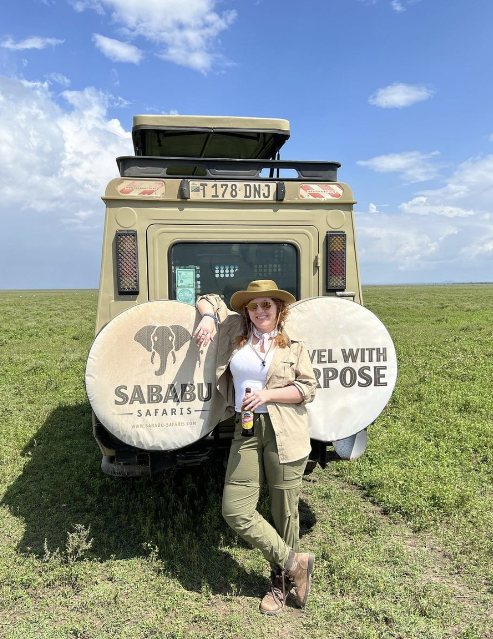 A woman wearing a hat, sunglasses, and safari gear leaning against a safari vehicle with promotional covers in an open grassy landscape under a blue sky with clouds.