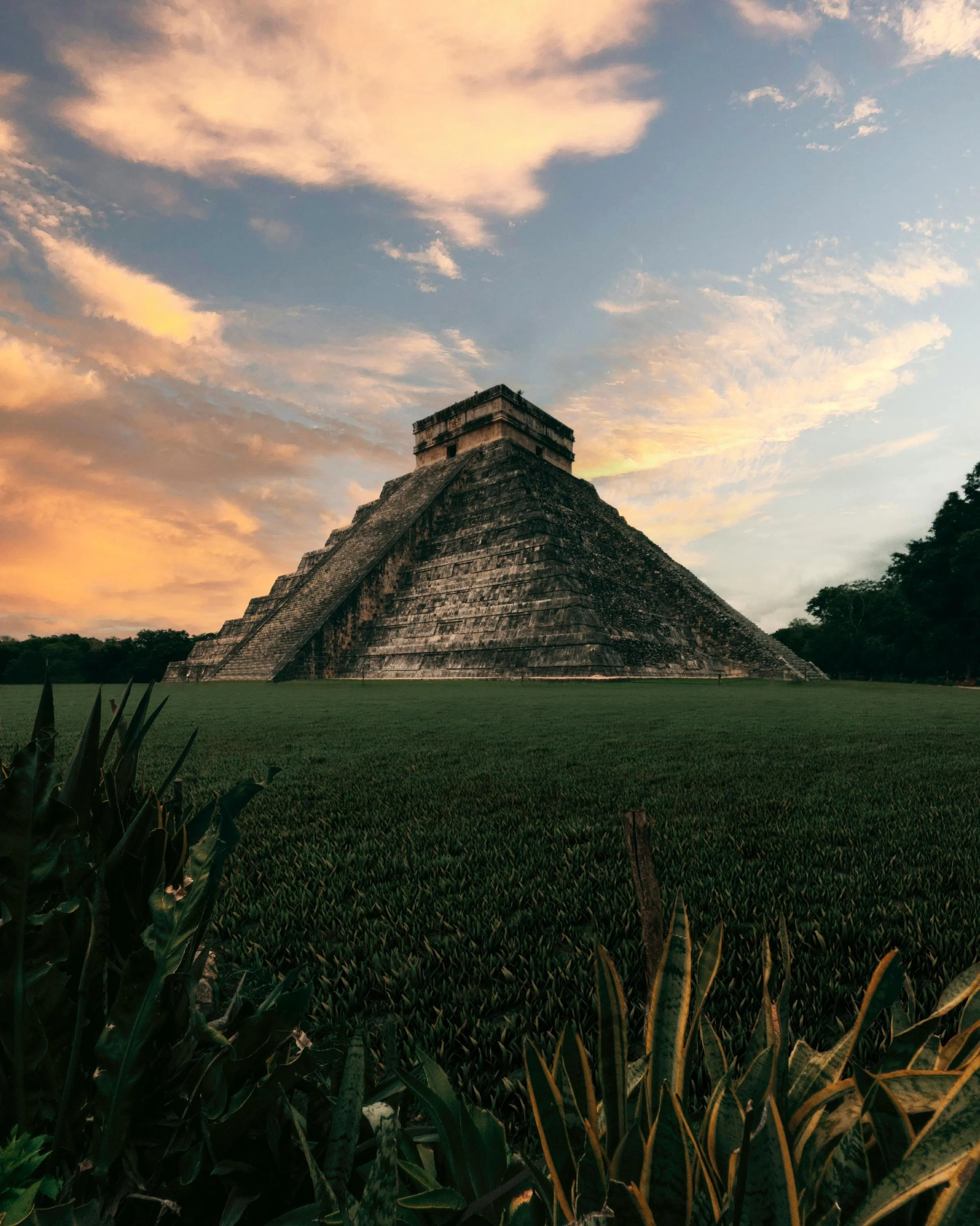 An ancient pyramid at sunrise or sunset, surrounded by green grass and trees, with colorful clouds in the sky.