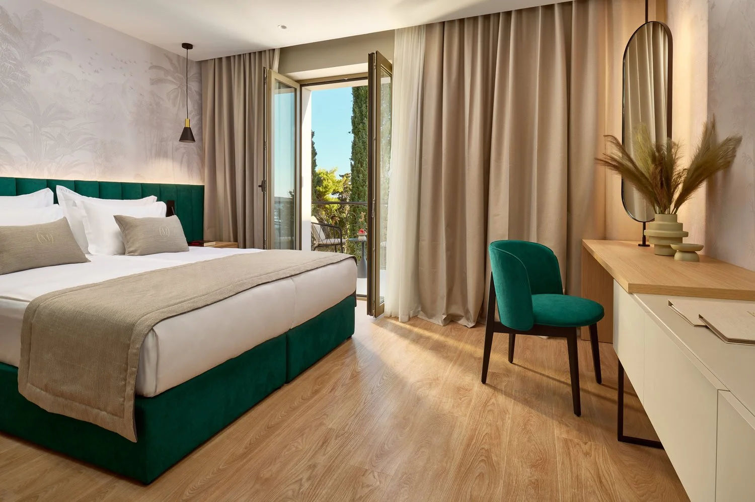 A bright, modern hotel room with a large bed with white linens and beige pillows, a balcony with outdoor seating, a vanity desk with a green chair, and decorative vases with dried grasses.