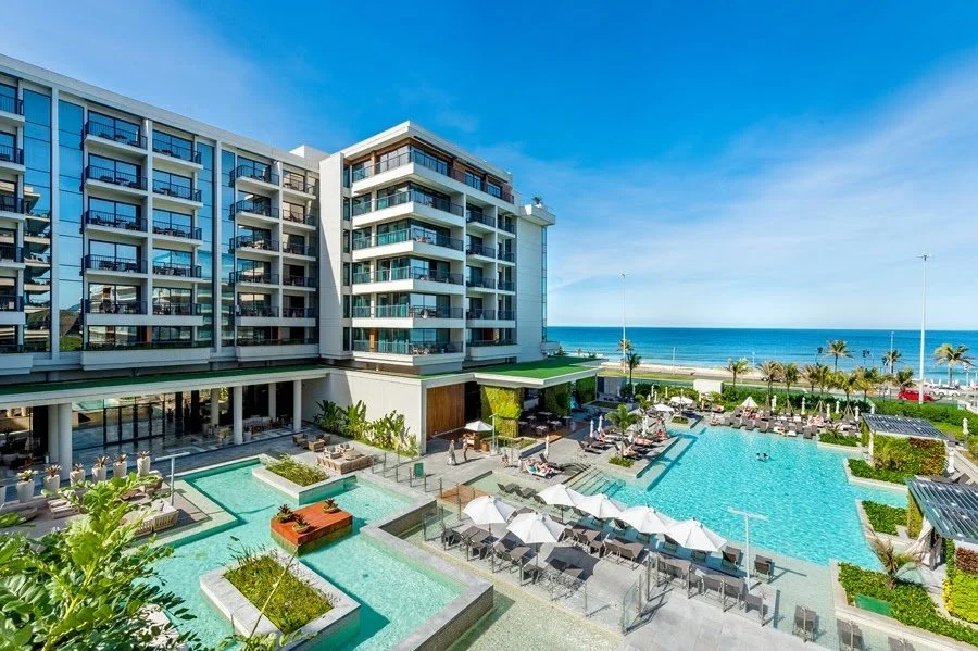Luxury hotel with multiple pools, outdoor lounge chairs, umbrellas, and a view of the ocean with palm trees.