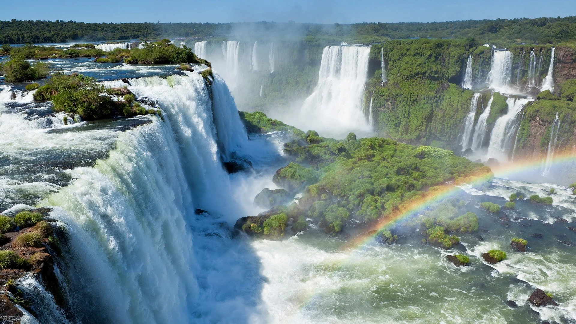 Multiple waterfalls cascading over cliffs with lush green vegetation, mist rising from the water, and a rainbow forming near the river's surface.